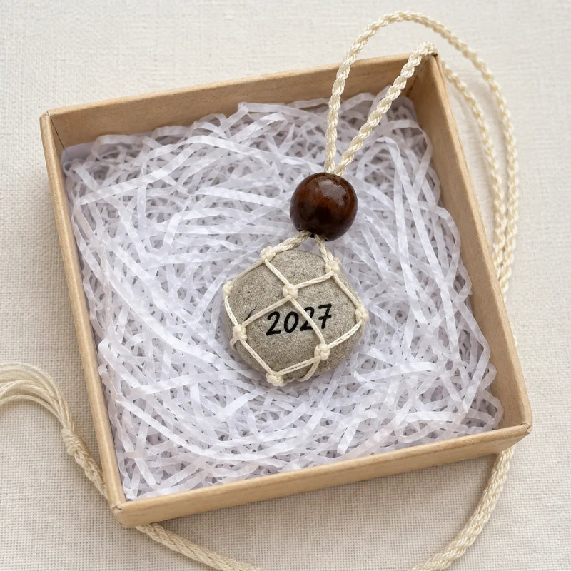A handmade pendant necklace featuring a gray stone with '2027' engraved on it, wrapped in beige string, hanging from a cream-colored cord with a dark brown wooden bead. The necklace is displayed inside a light brown gift box filled with white paper shred.