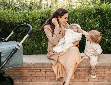 Woman sitting on a wall with two children, one in her arms and another standing next to her, with a stroller nearby.
