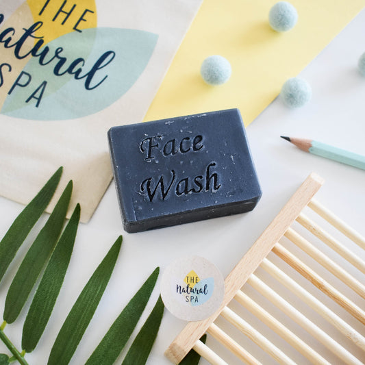 Bar of face wash with 'The Natural Spa' branding on a white surface with green leaves and a wooden soap dish.