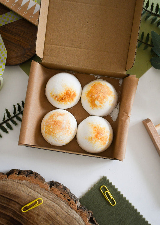 Four bath bombs with orange specks in a cardboard box on a white surface with decorative elements.