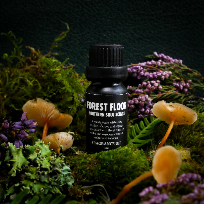 Bottle of 'Forest Floor' fragrance oil surrounded by mushrooms and purple flowers on a dark background