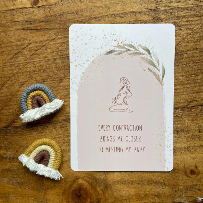 Card with a pregnant woman illustration and text on a wooden surface with two small crochet items.