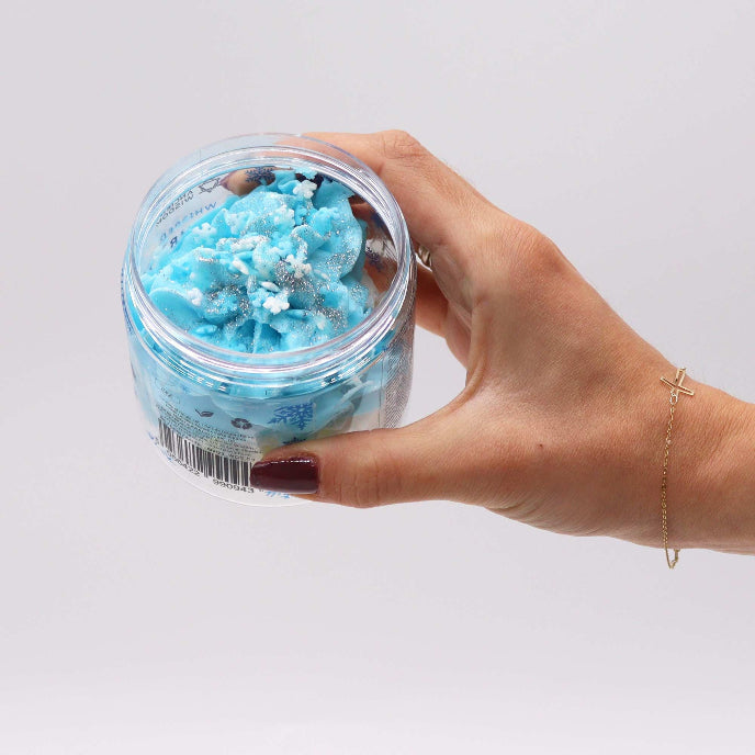 Hand holding a jar of blue bath salt scrub against a light gray background
