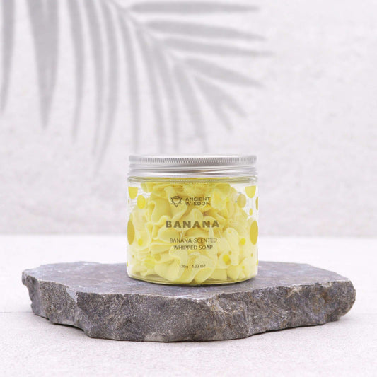 Jar of banana-flavored whipped soap on a stone platform with a white background