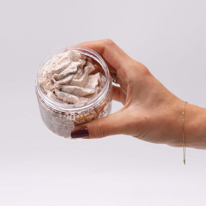 Hand holding a jar of grayish bath salt scrub against a light background