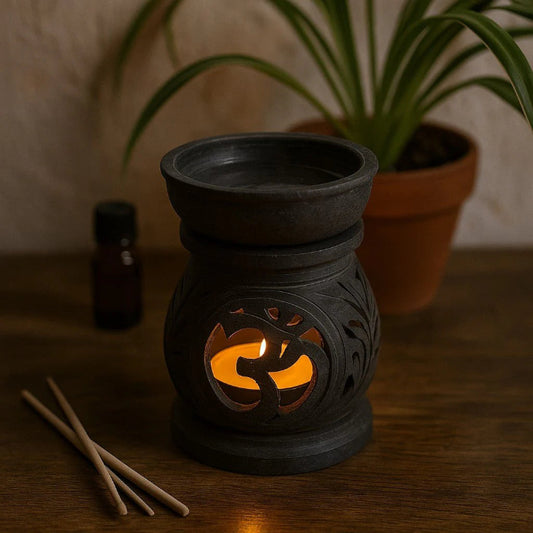 Black ceramic oil burner with a lit candle inside, placed on a wooden surface with a plant in the background.