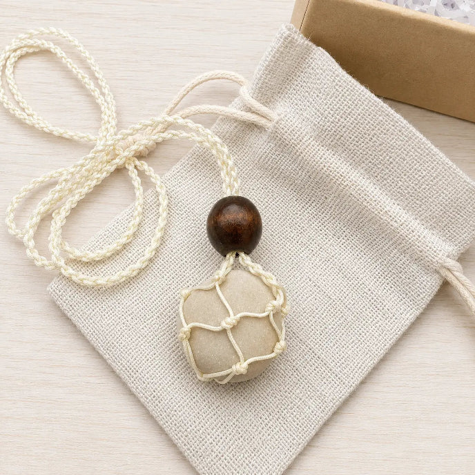 A handmade pendant necklace featuring a cream-colored woven net bag with a dark brown wooden bead at the top, resting on a matching cream drawstring pouch.