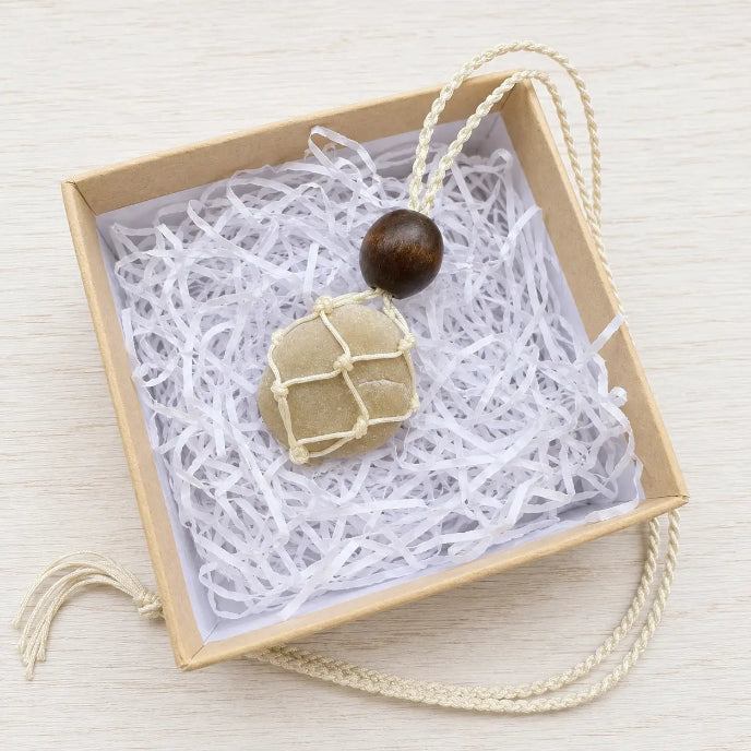 A small beige fabric pouch with cream cord netting and a dark brown wooden bead, presented inside a square kraft gift box filled with white paper shred on a light wood surface.