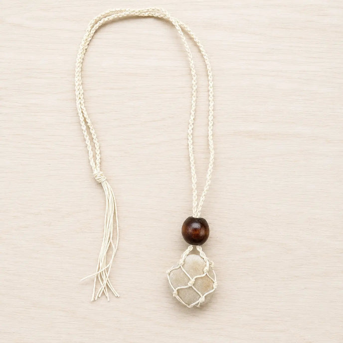 A bohemian-style pendant necklace featuring a cream braided cord, a dark brown wooden bead, and a white netted pouch containing a small beige stone, displayed on a light wood background.