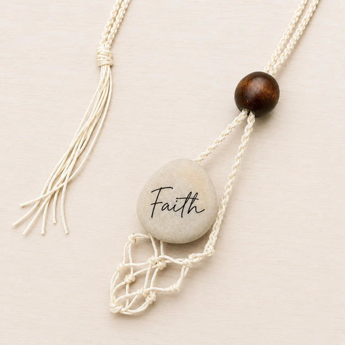 A bohemian-style macrame necklace featuring a light gray stone engraved with the word 'Faith', a dark brown wooden bead, and cream-colored cords with tassels, displayed on a light beige background.