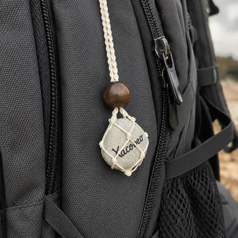 A close-up of a black backpack with a woven cream cord charm holding a small gray stone engraved with the word 'Vacero', with a dark brown wooden bead above it.