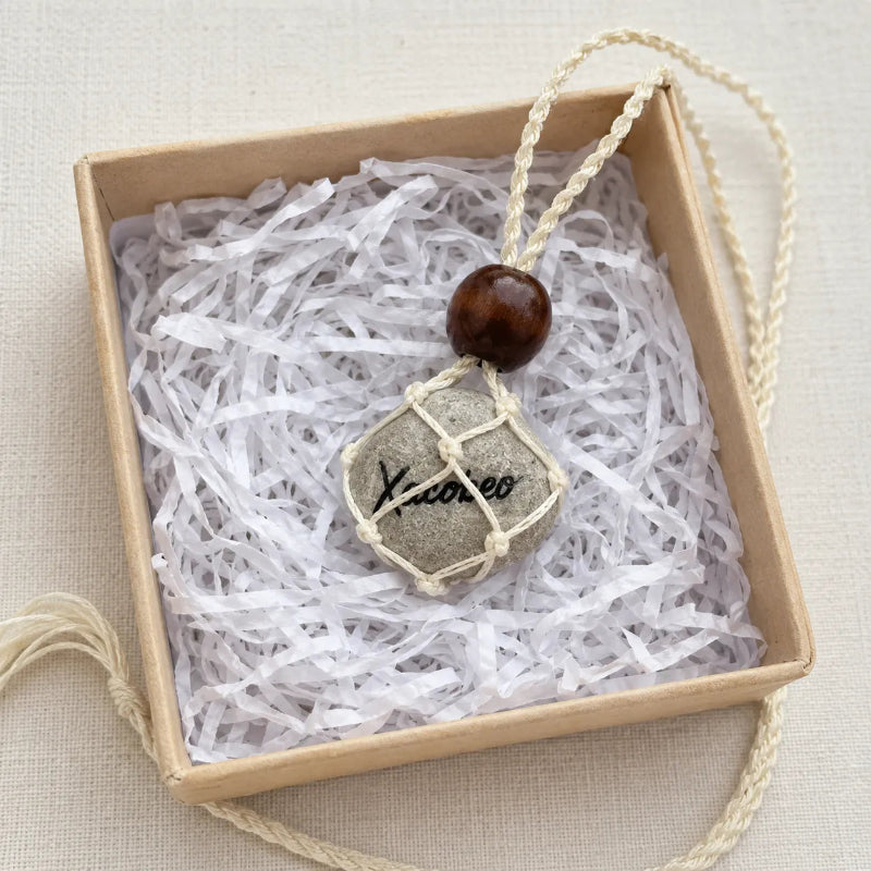 A handmade pendant necklace featuring a small gray stone with black script, wrapped in cream cord, and topped with a dark brown wooden bead, presented inside a kraft gift box with white paper shred.
